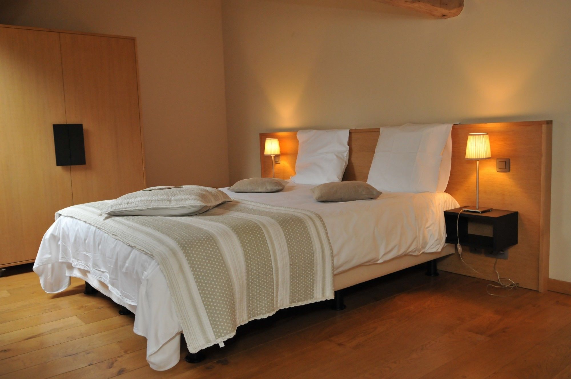 Een net opgemaakt bed met een wit en beige stippenbedekking, twee bedlampjes en een houten nachtkastje.
