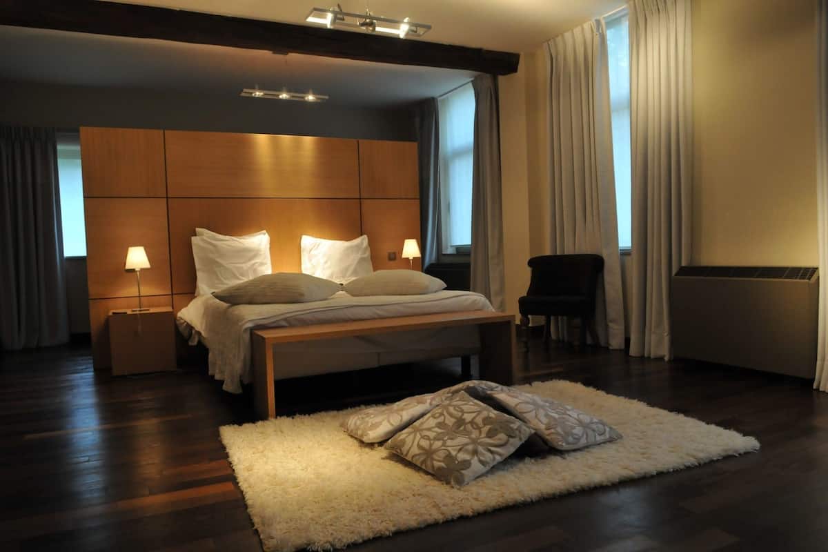 A modern bedroom with a wooden headboard, a large bed with white linens, two bedside lamps, a fluffy rug, and a chair.