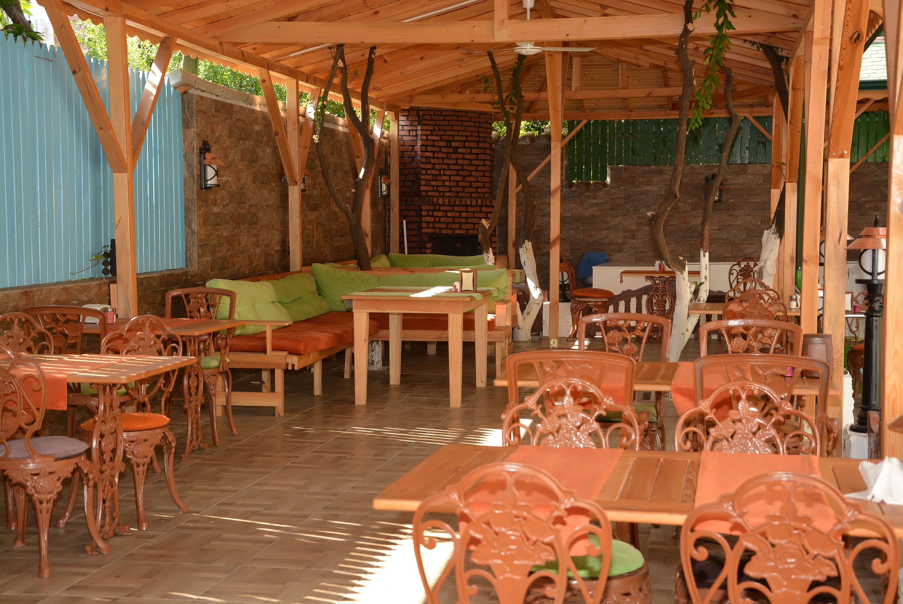 An outdoor dining area with wooden tables and chairs, a brick wall, and a covered seating area with green cushions.