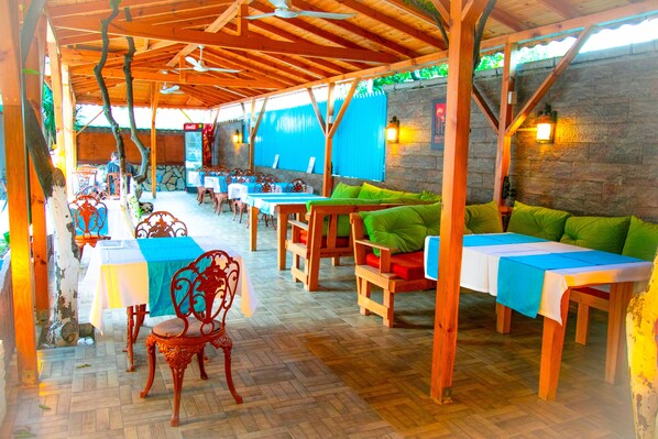 An outdoor patio area with wooden tables and chairs, stone walls, and a blue curtain backdrop. - Kaliptus Hotel (Kemer)