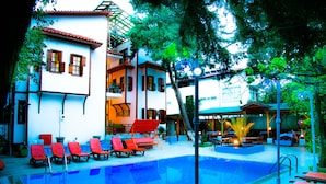 Una piscina con sillas reclinables rojas, una zona de estar cubierta y un edificio con varias ventanas y balcones.