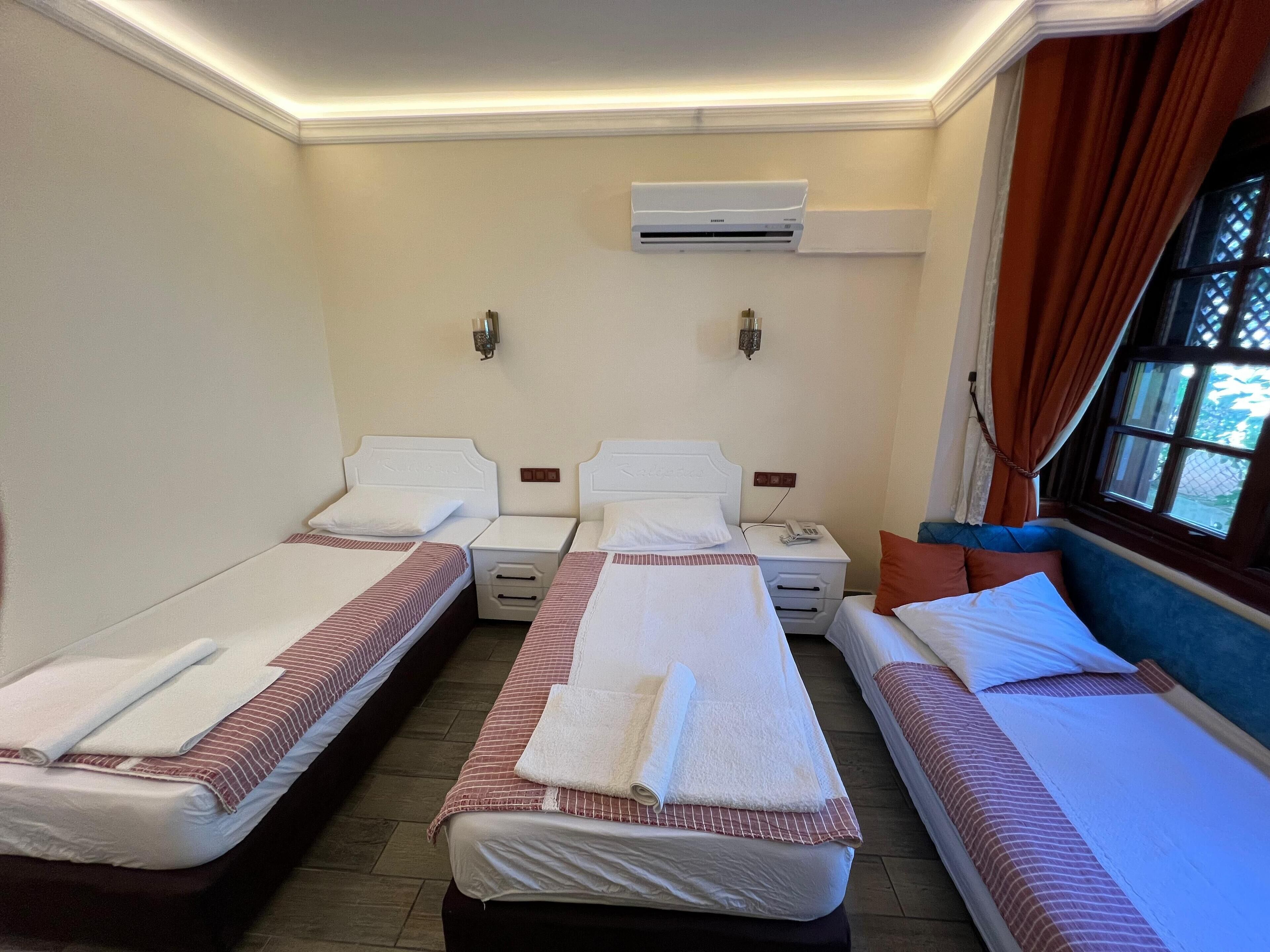A hotel room with three single beds, each with a white headboard, a small bedside table, and a lamp. There is a window with curtains on the right side.