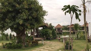 A pathway leading to a series of wooden chalets with thatched roofs, surrounded by lush greenery and a large tree.
