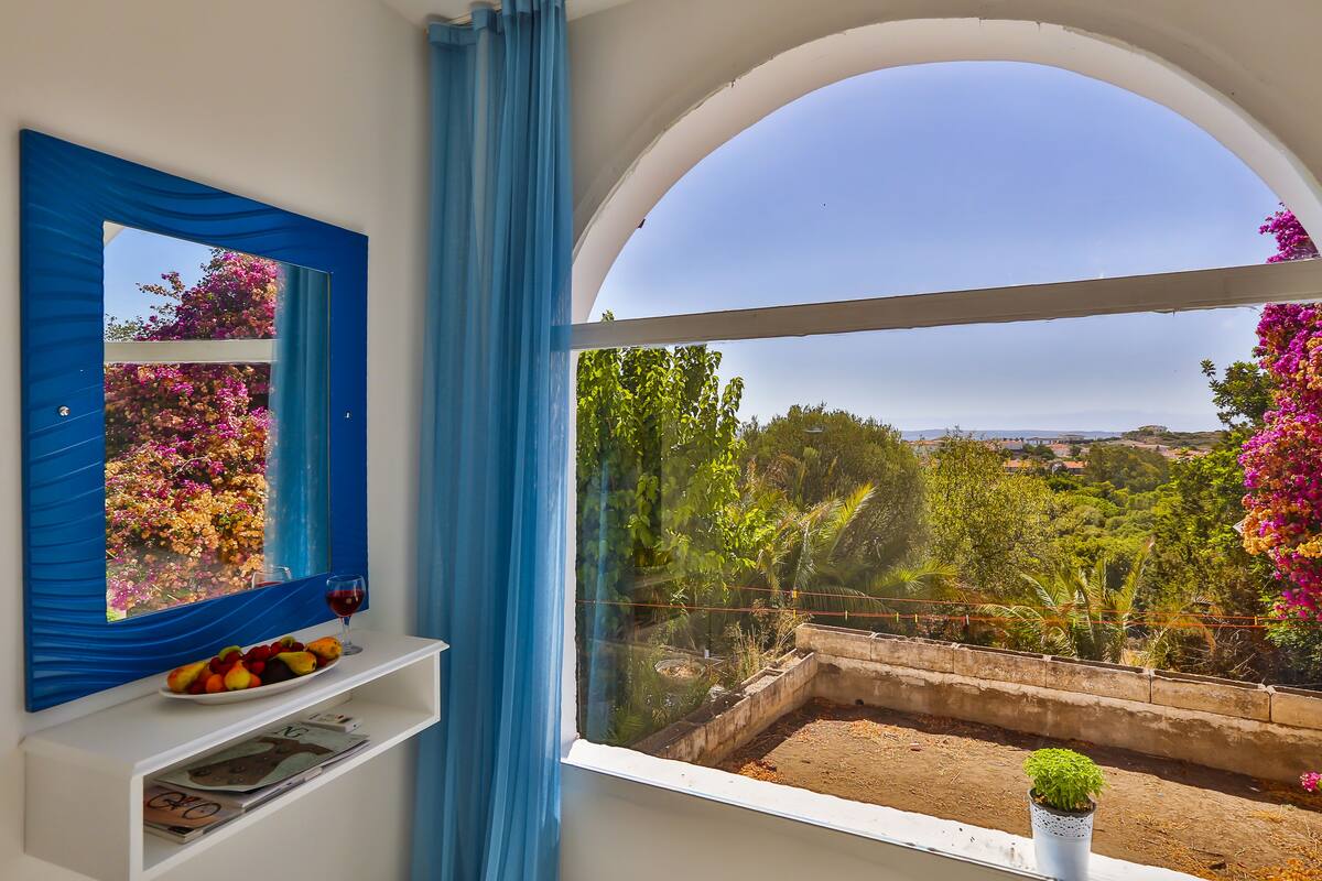 A room with a large arched window, a blue-framed mirror, a white shelf with a fruit bowl and a glass of red wine, and a small potted plant.