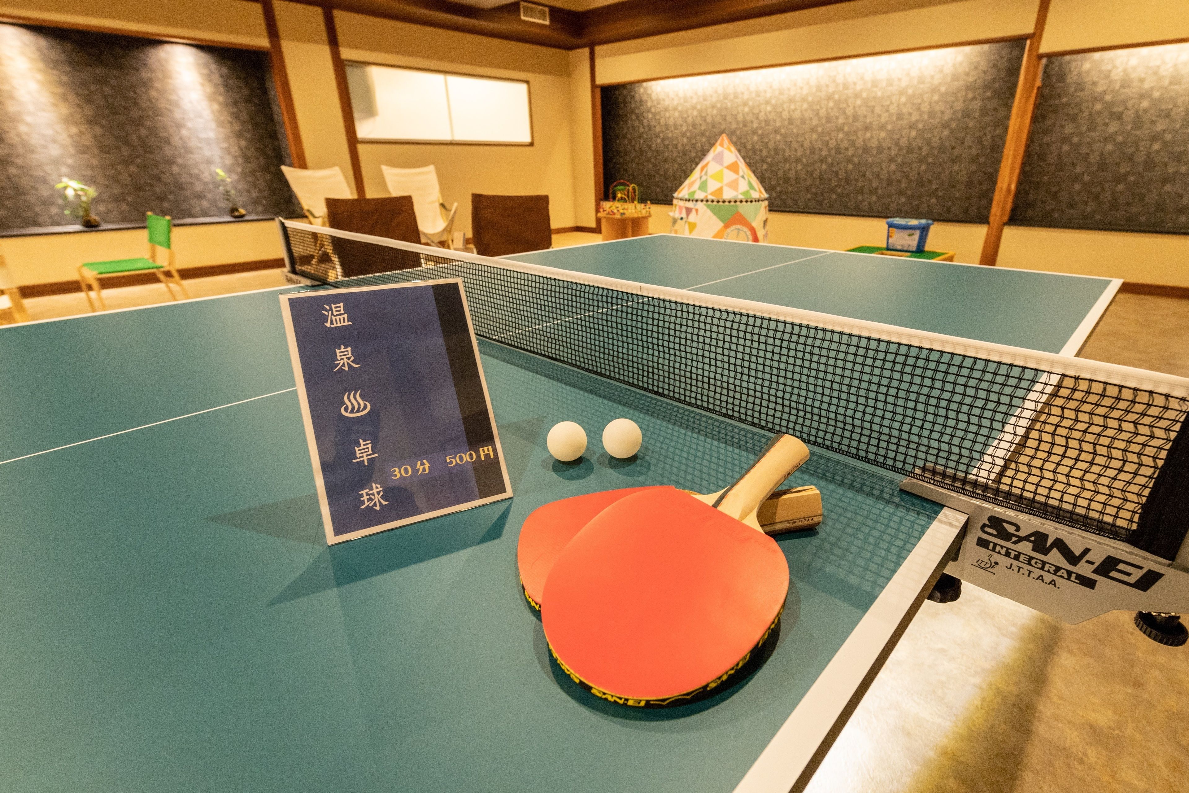 A ping pong table with two paddles and two balls, a sign with pricing, and a small house decoration.