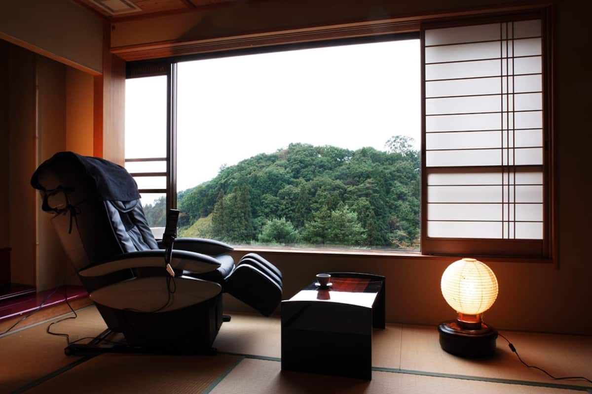 A room with tatami flooring, a futon bed, a desk with a lamp, and a view of a forest through a window.