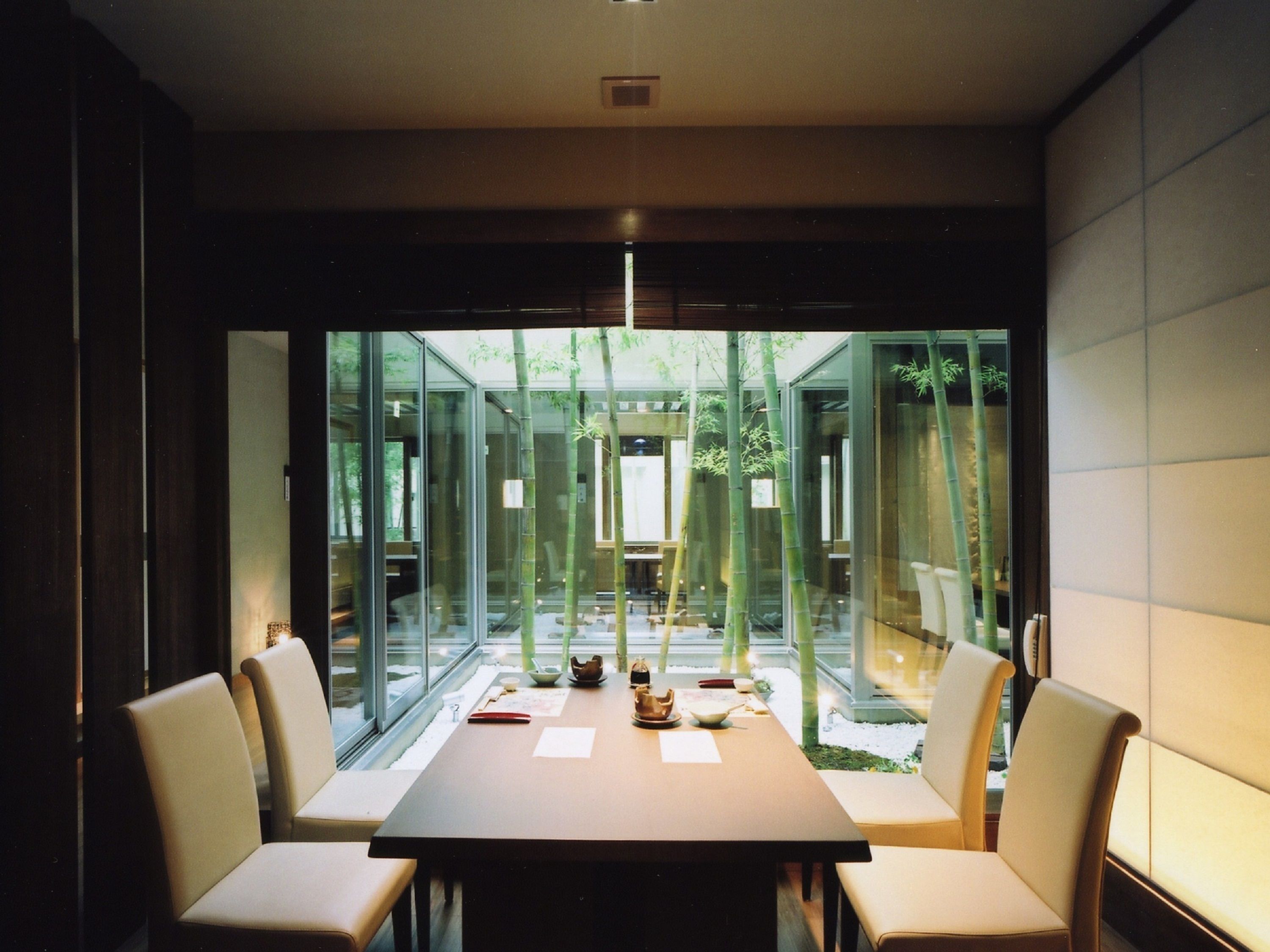 A modern dining area with a long table and six chairs, large windows, and bamboo plants.
