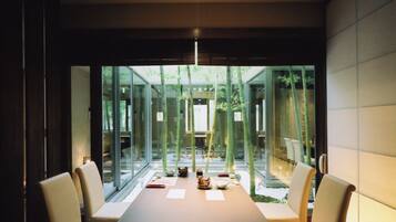 A modern dining area with a long table and six chairs, large windows, and bamboo plants.
