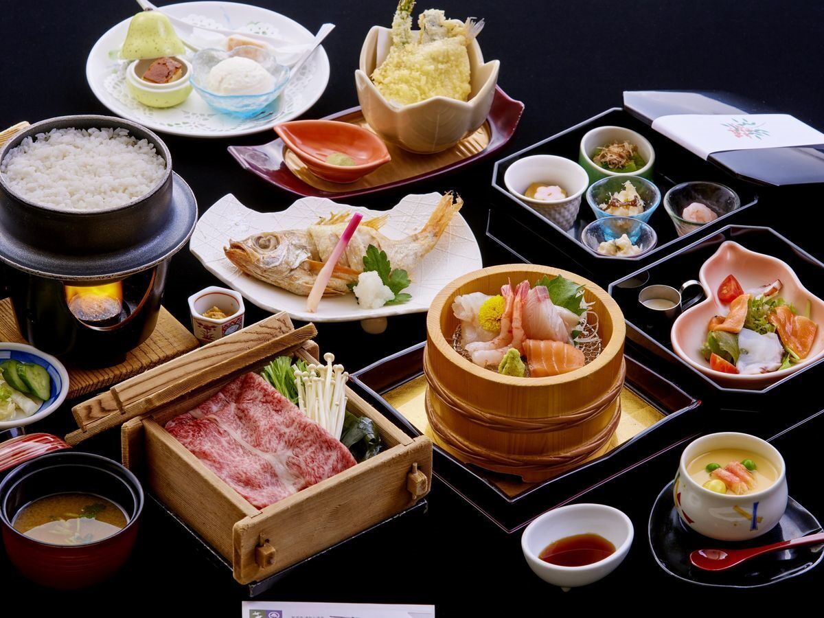 A variety of Japanese dishes including sushi, sashimi, and tempura, served on a black table with multiple small dishes and a hot pot.