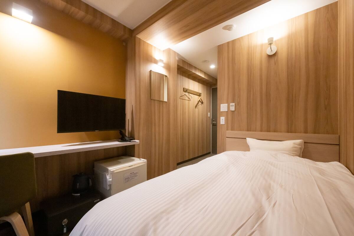 A hotel room with a bed, a television mounted on the wall, a small desk with a kettle, and wooden paneling on the walls.