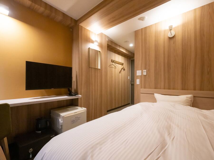 A hotel room with a bed, a television mounted on the wall, a small desk with a kettle, and wooden paneling on the walls.