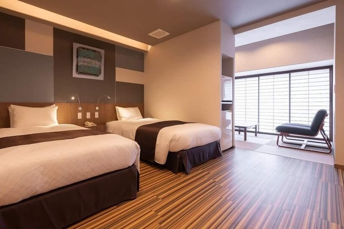 A hotel room with two beds, a wooden floor, a large window with blinds, and a chair by the window.