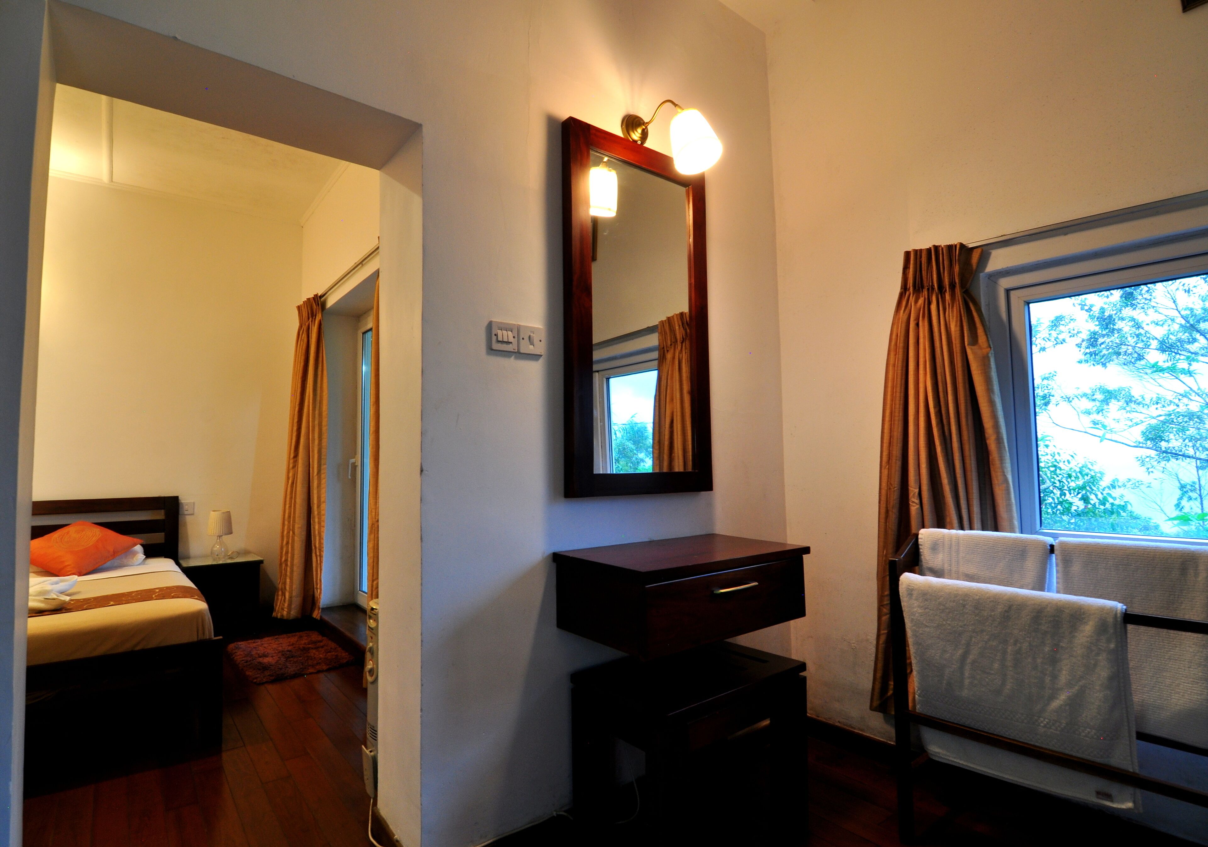 A hotel room with a bed, a bedside table, a mirror, a window with curtains, and a view of trees outside.