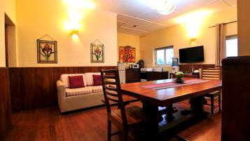 A dining area with a wooden table and chairs, a sofa with cushions, a television, and artwork on the walls.