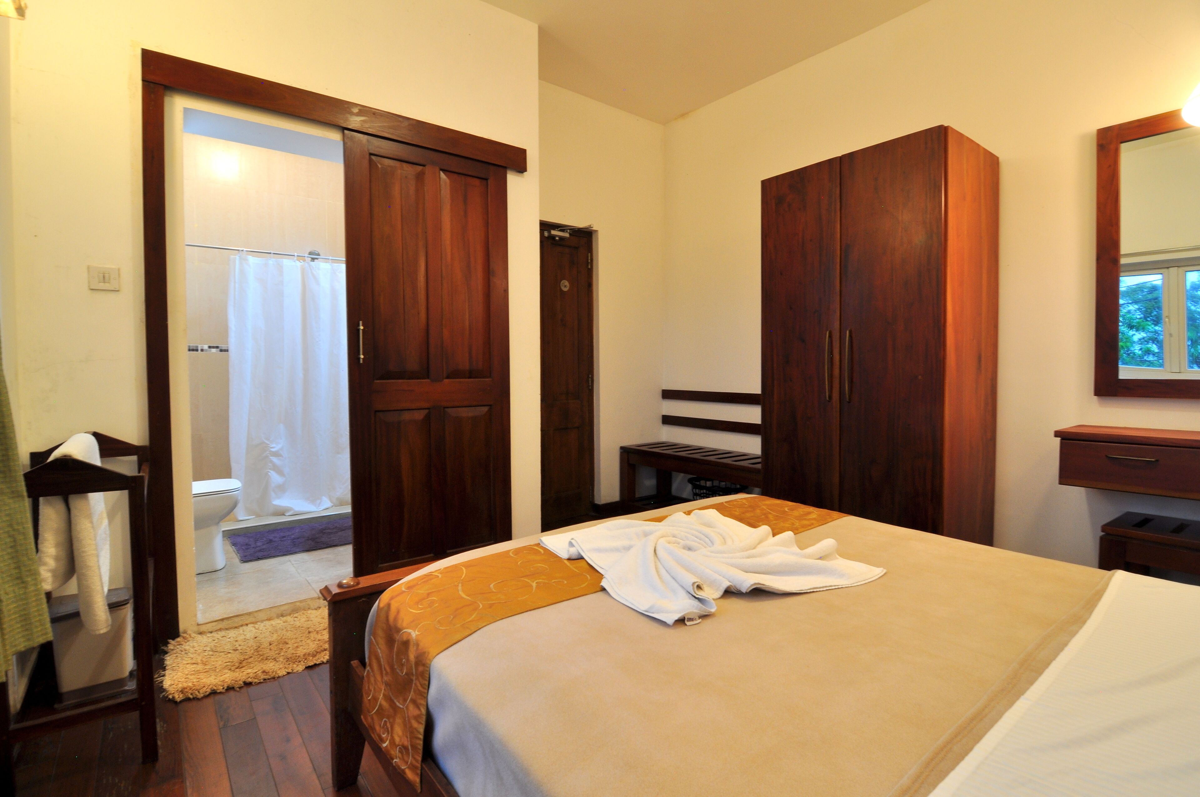 A hotel room with a bed, wooden wardrobe, a bathroom with a shower curtain, and a mirror on the wall.
