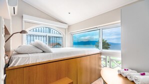 A bedroom with a wooden bed, a bench, a table with towels, and a view of the sea through large windows.