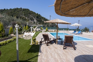 Uma área à beira da piscina com duas espreguiçadeiras, uma mesinha e um guarda-sol de palha.