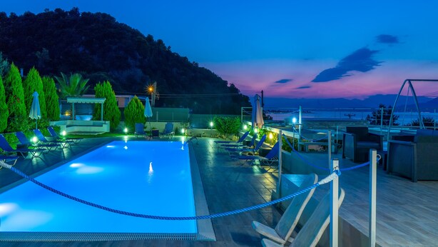 A swimming pool with lounge chairs and umbrellas, surrounded by a wooden deck and a view of the sea and hills.