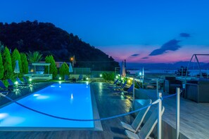 A swimming pool with lounge chairs and umbrellas, surrounded by a wooden deck and a view of the sea and hills.