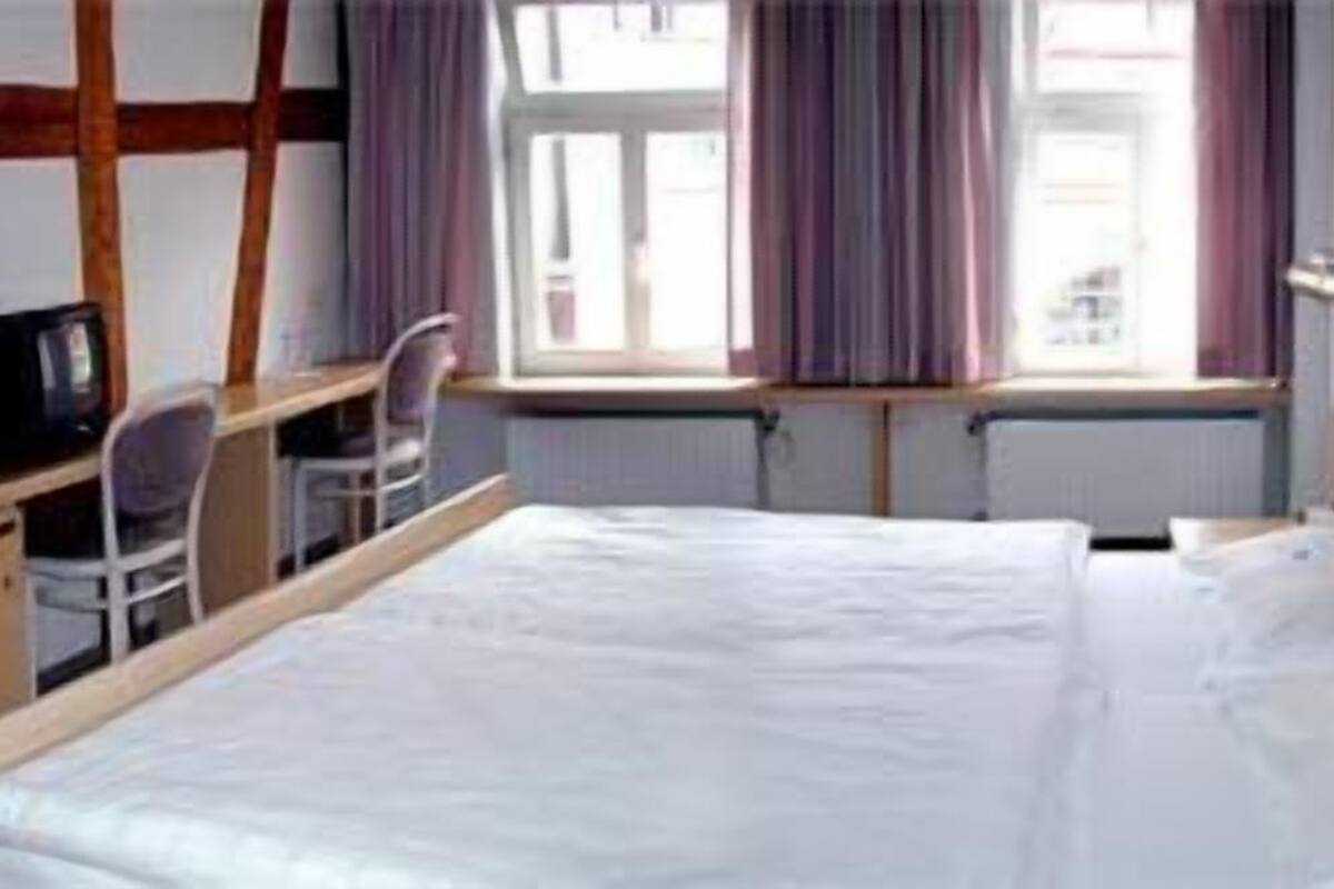 Una habitación de hotel con cama, escritorio, silla y ventana con cortinas.