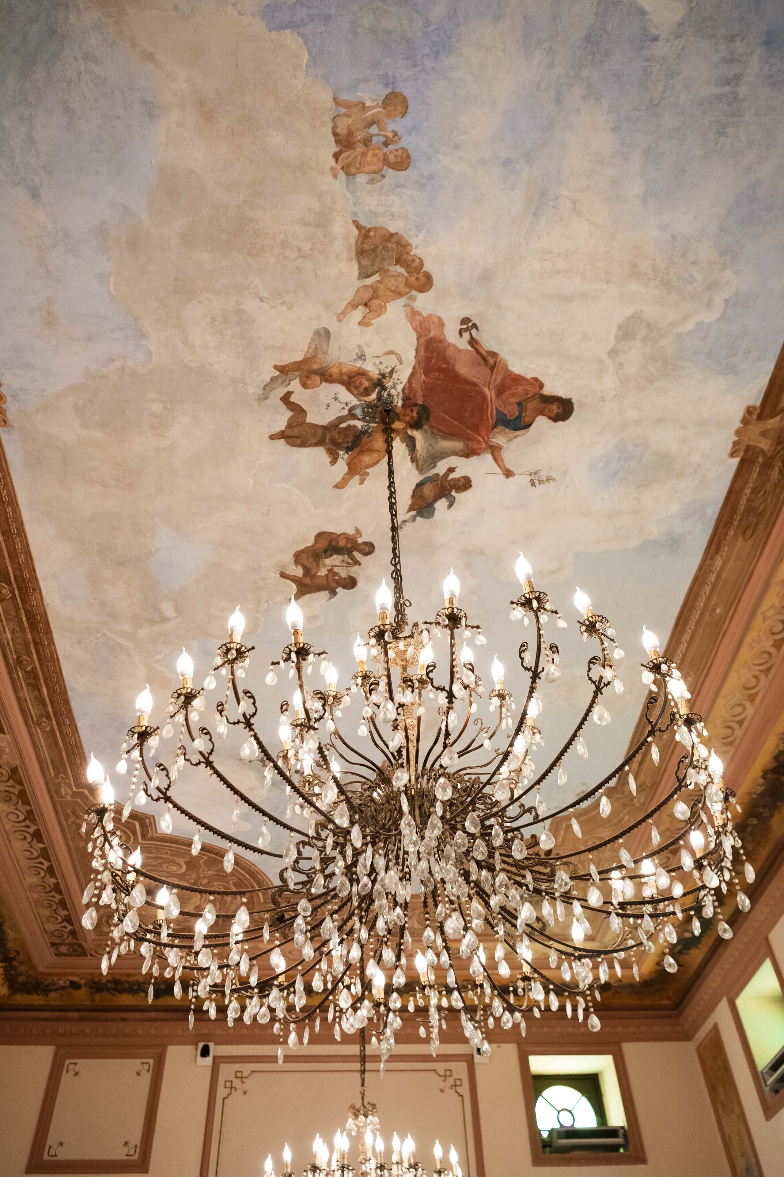 A grand chandelier with multiple arms and crystal decorations, set against a ceiling with painted clouds and cherubs.