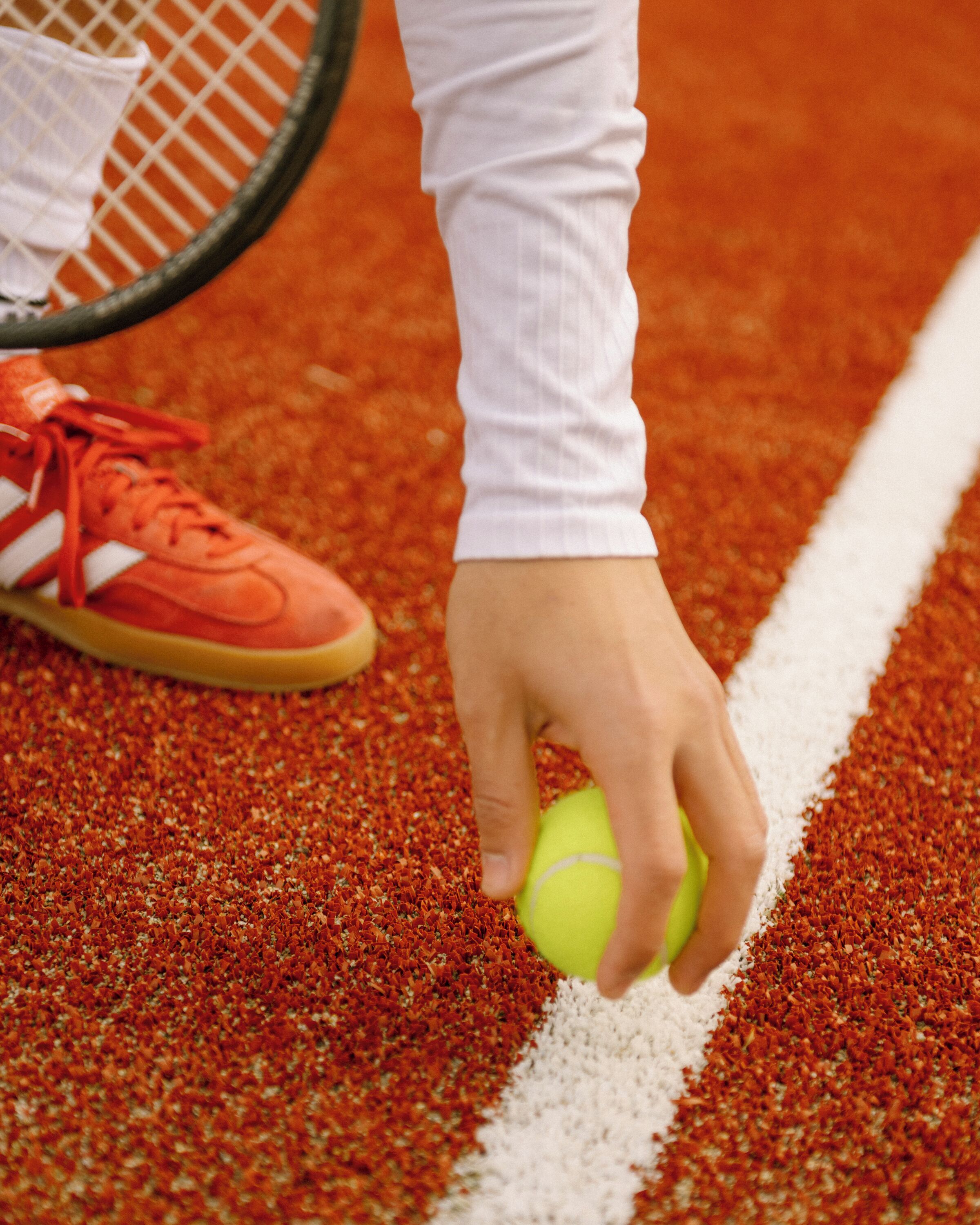 Una mano sosteniendo una pelota de tenis en una cancha de arcilla roja con líneas blancas, y una raqueta de tenis y zapatos rojos al fondo.
