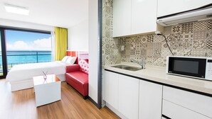 Une chambre d’hôtel moderne avec un lit, un canapé rouge, un coin cuisine équipé d’un four à micro-ondes et une vue sur la mer.