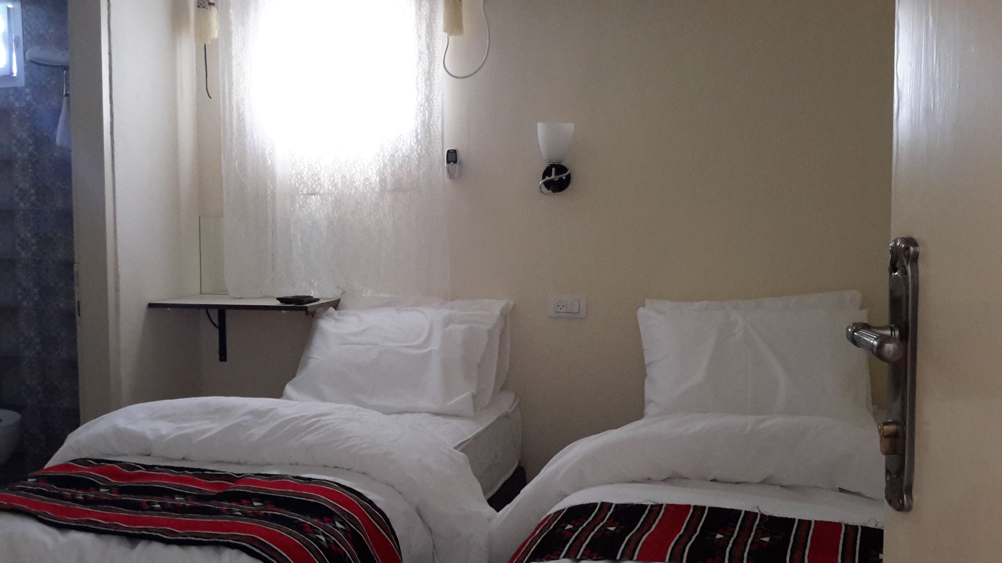 A small, simple room with two beds, a small table, and a wall-mounted light.