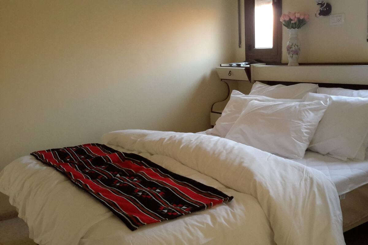 Cama com roupa de cama branca e um cobertor vermelho e preto, um vaso com flores na mesinha de cabeceira e uma janela com cortinas.