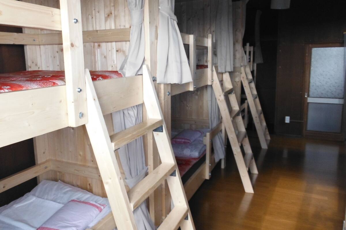 Kerrossängyt, joissa on puinen runko ja valkoiset vuodevaatteet, on asetettu riviin huoneeseen, jossa on puulattia ja taustalla näkyy ovi.