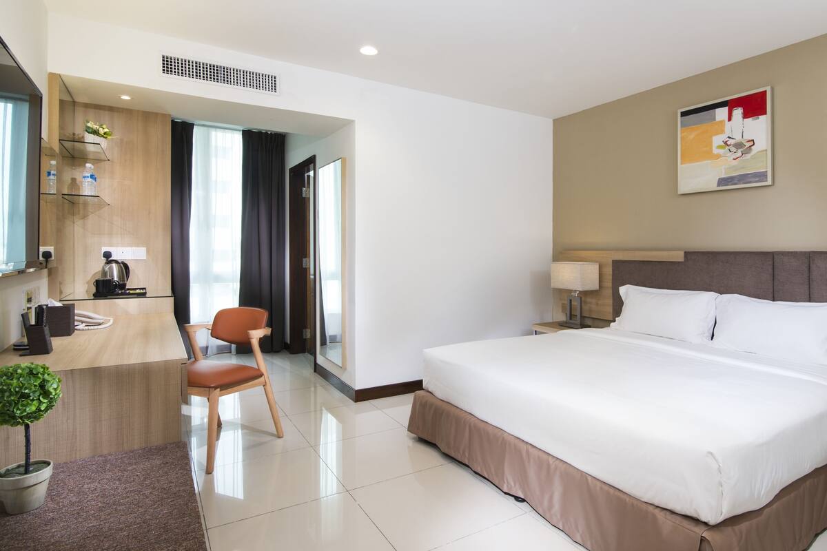 Habitación de hotel moderna con una cama grande, un escritorio con cafetera, una silla y una planta pequeña.