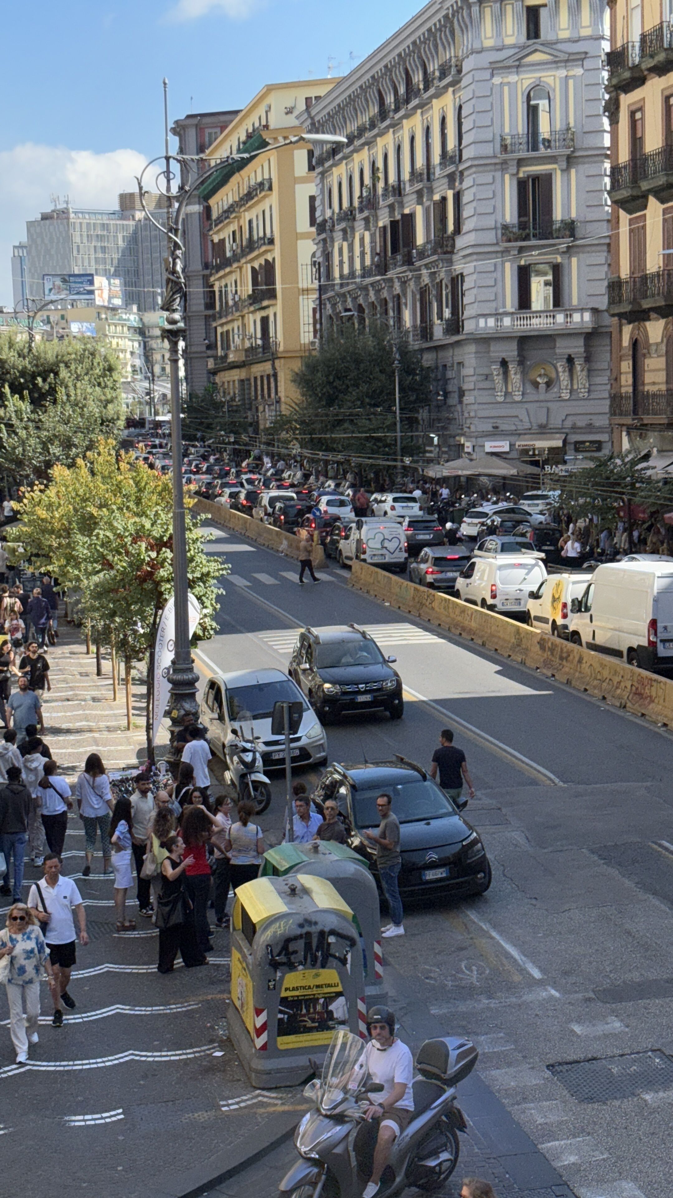 Uma rua movimentada no centro da cidade, com pedestres, carros estacionados e um motociclista.