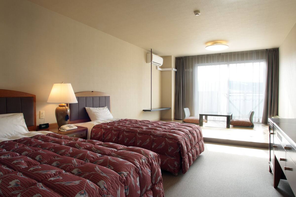 Een hotelkamer met twee bedden, een bureau, een lamp en uitzicht op de stad.