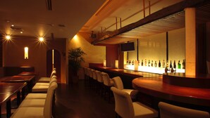 A bar area with wooden tables and white cushioned chairs, a long counter with bottles, and a TV mounted on the wall.