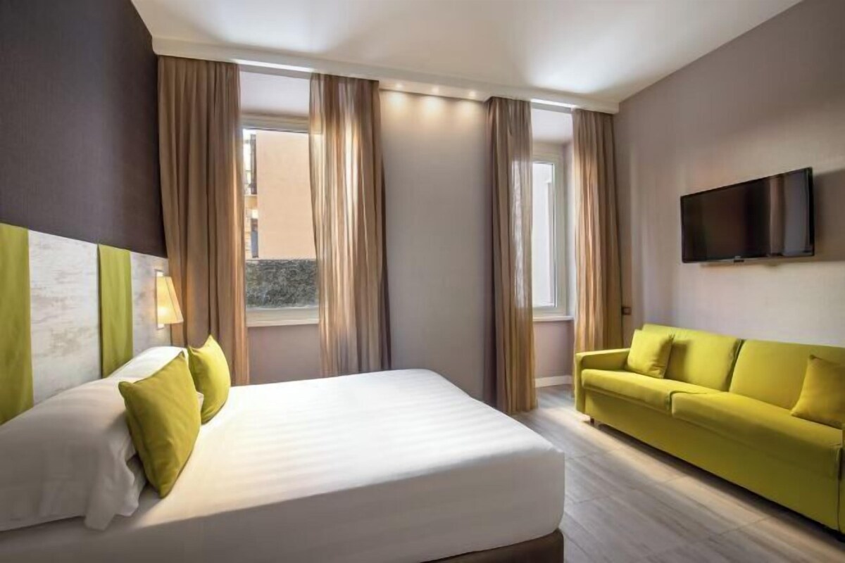 Habitación de hotel con cama, sofá, televisión y ventanas grandes con cortinas.