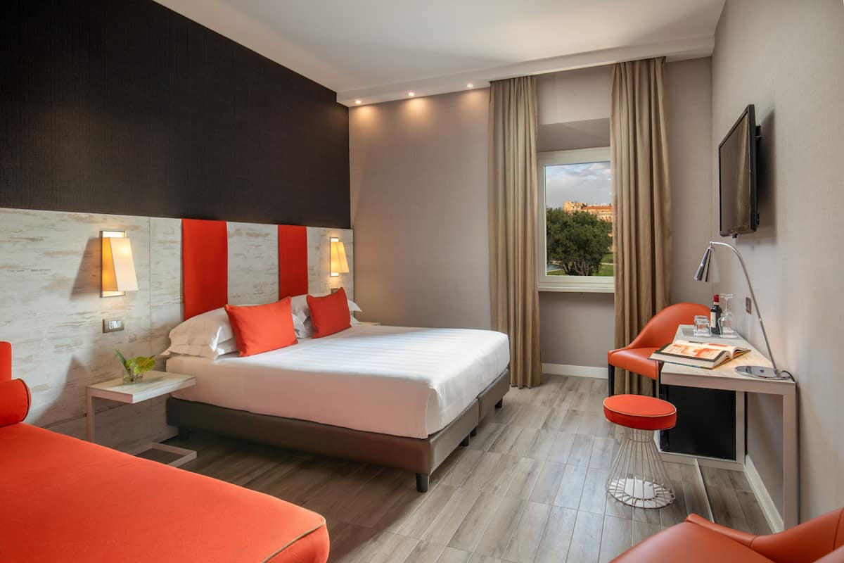 A modern hotel room with a large bed, a desk with a chair, a TV mounted on the wall, and a window with curtains.