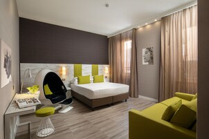 A modern hotel room with a large bed, a desk with a chair, a sofa, and a large window with curtains.