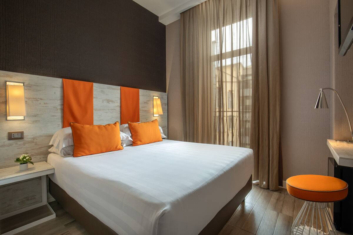 A hotel room with a large bed, two bedside lamps, a nightstand with a flower, and a modern bathroom with a mirror and sink.
