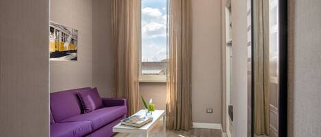A modern hotel room with a purple sofa, a small white table with a book and a plant, and a large window with curtains.
