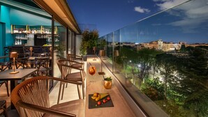 A modern outdoor dining area with tables and chairs, a bar with shelves of bottles, and a view of a cityscape and river. - The Guardian Hotel (Rome)
