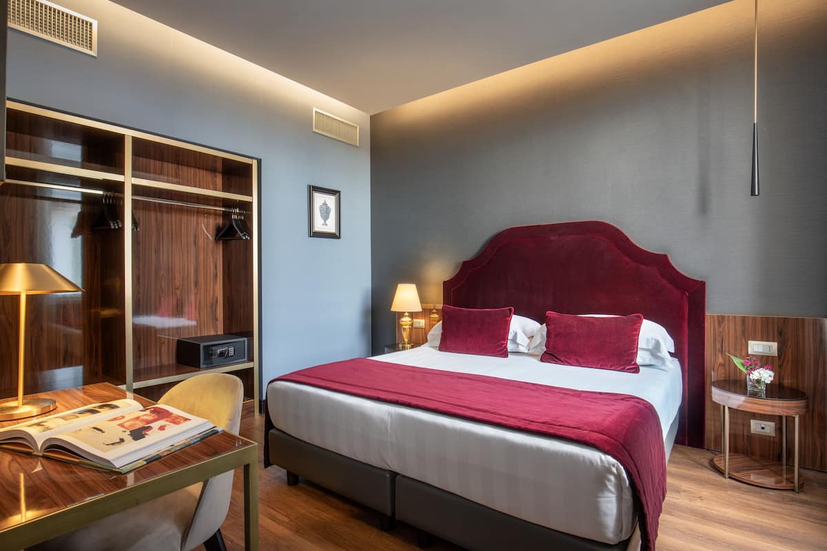 A hotel room with a large bed, a desk with a lamp, a wardrobe, and a bedside table with a vase.