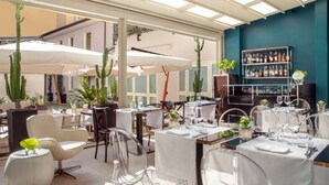 An outdoor dining area with clear chairs, white tablecloths, and umbrellas. There are potted plants and a bar area with a wine rack. - The Guardian Hotel (Rome)