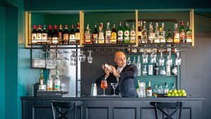 A bar area with shelves of liquor bottles and glasses, a bartender preparing a drink, and two black chairs in front. - The Guardian Hotel (Rome)