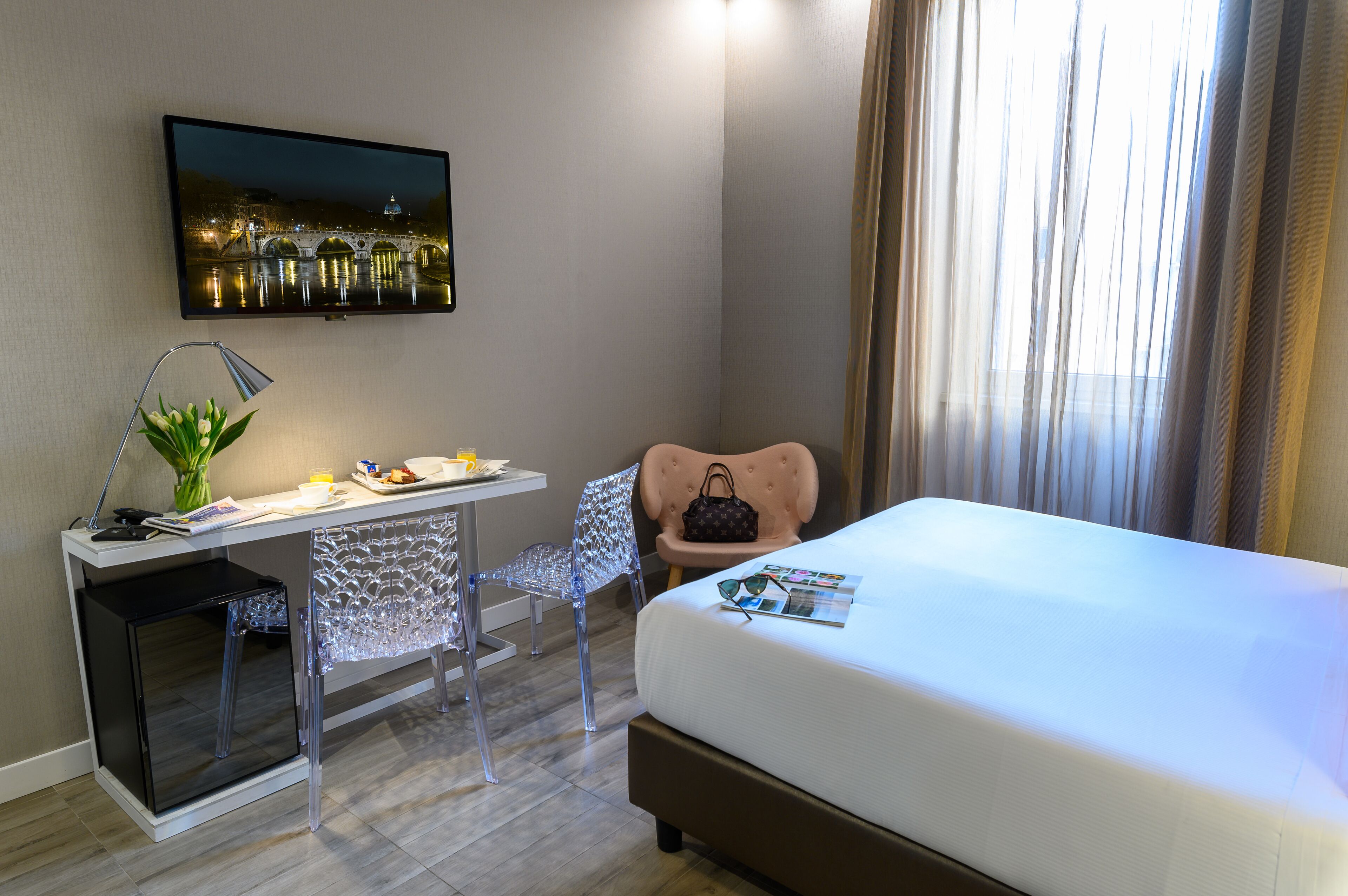 Habitación de hotel con cama, escritorio con computadora, silla, televisor en la pared y ventana con cortinas.
