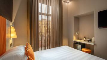 A hotel room with a bed, a desk with a chair, a TV mounted on the wall, and a window with curtains.