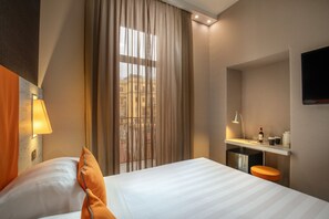 A hotel room with a bed, a desk with a chair, a TV mounted on the wall, and a window with curtains.