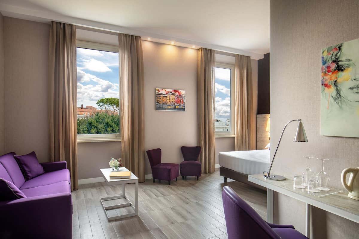 A modern hotel room with a large window, a bed, a desk with a lamp, and purple seating.