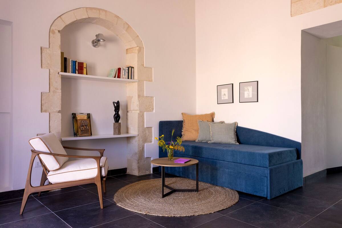 Uma sala de estar com um sofá azul, uma cadeira de madeira, uma mesinha redonda com um vaso e uma estante sob um arco.