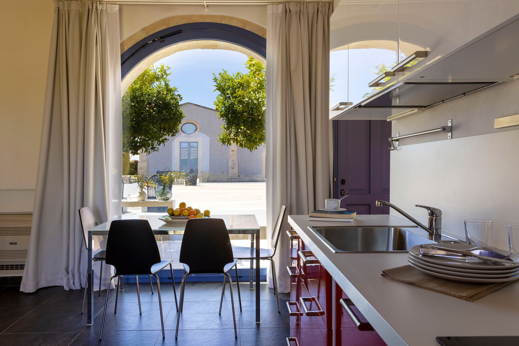 A modern kitchen with a dining area, featuring a large window with curtains, a sink, and a dining table with chairs.