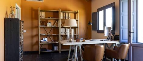 A cozy room with a wooden ceiling, a desk with chairs, a bookshelf, and a window with blinds.